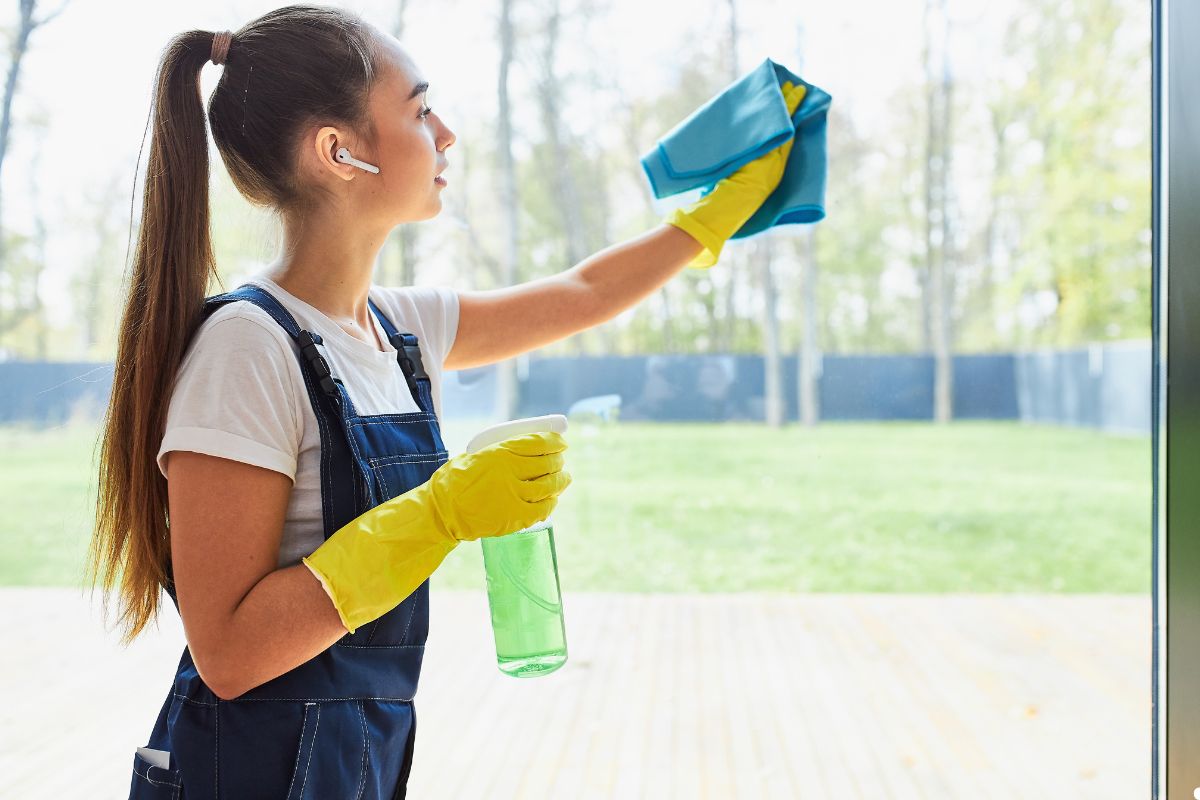 A woman wearing overalls and rubber gloves is cleaning a window with a cloth, focused on her task.